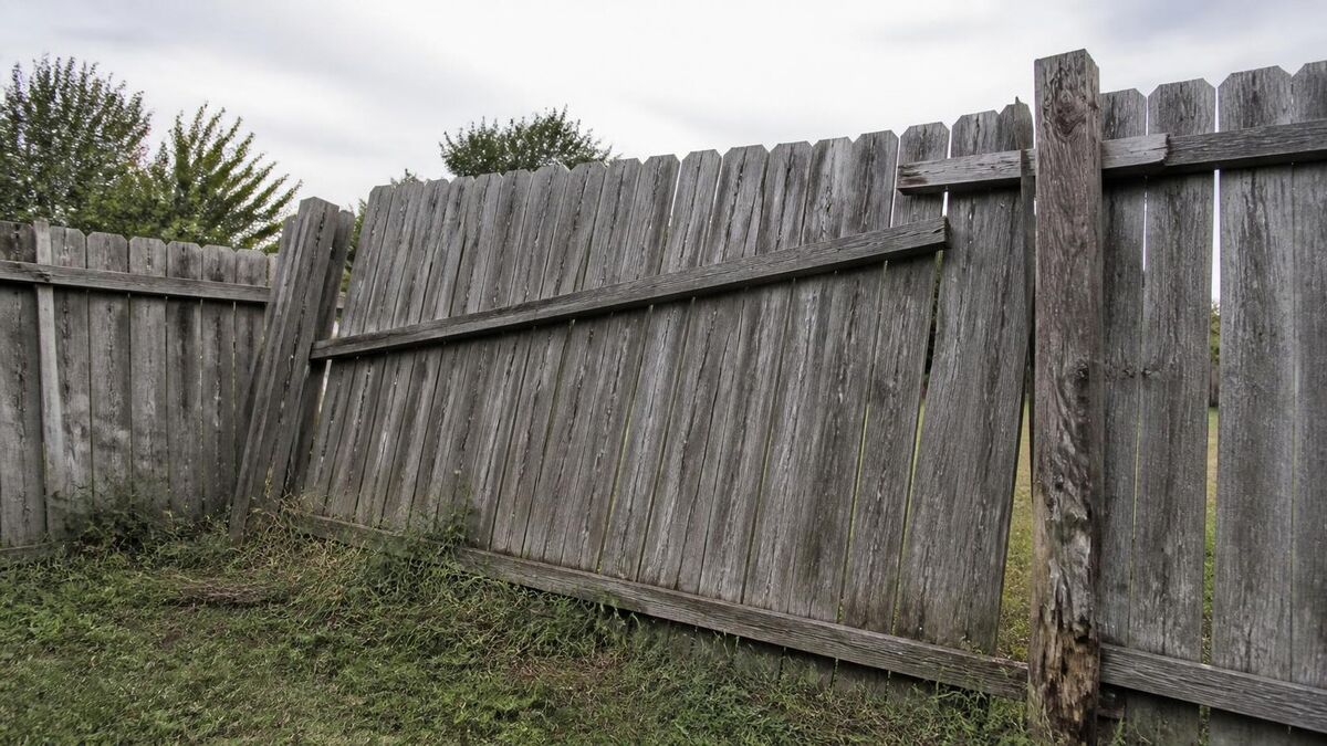 An old gray wood privacy fence leaning out of plumb, with a tilted post and a loose rail along the bottom